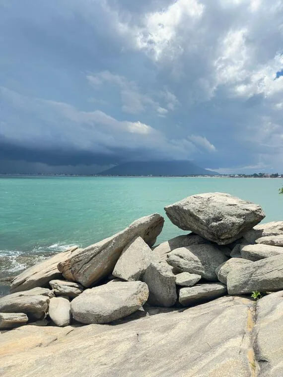 Pedras à beira do mar esverdeado em Rio das Ostras sob nuvens de chuva