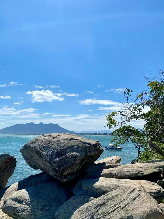 Pedra equilibrada no costão rochoso de Rio das Ostras com lancha e serra ao fundo